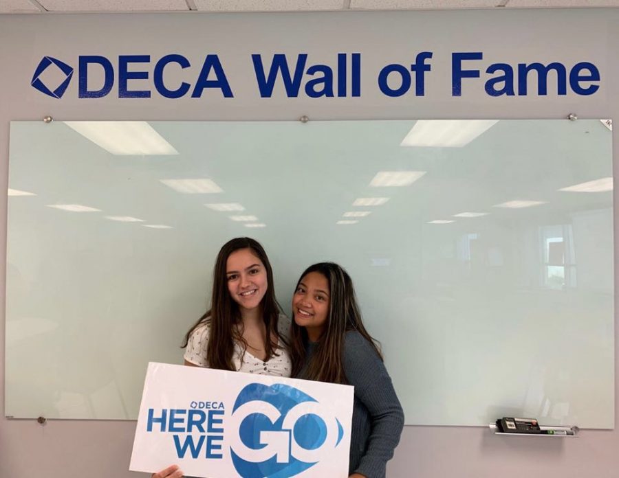 Seniors and DECAlicious 2019-2020 CEOs Caroline Schapmann and Meagan Mercado standing in front of the DECA Wall of Fame.