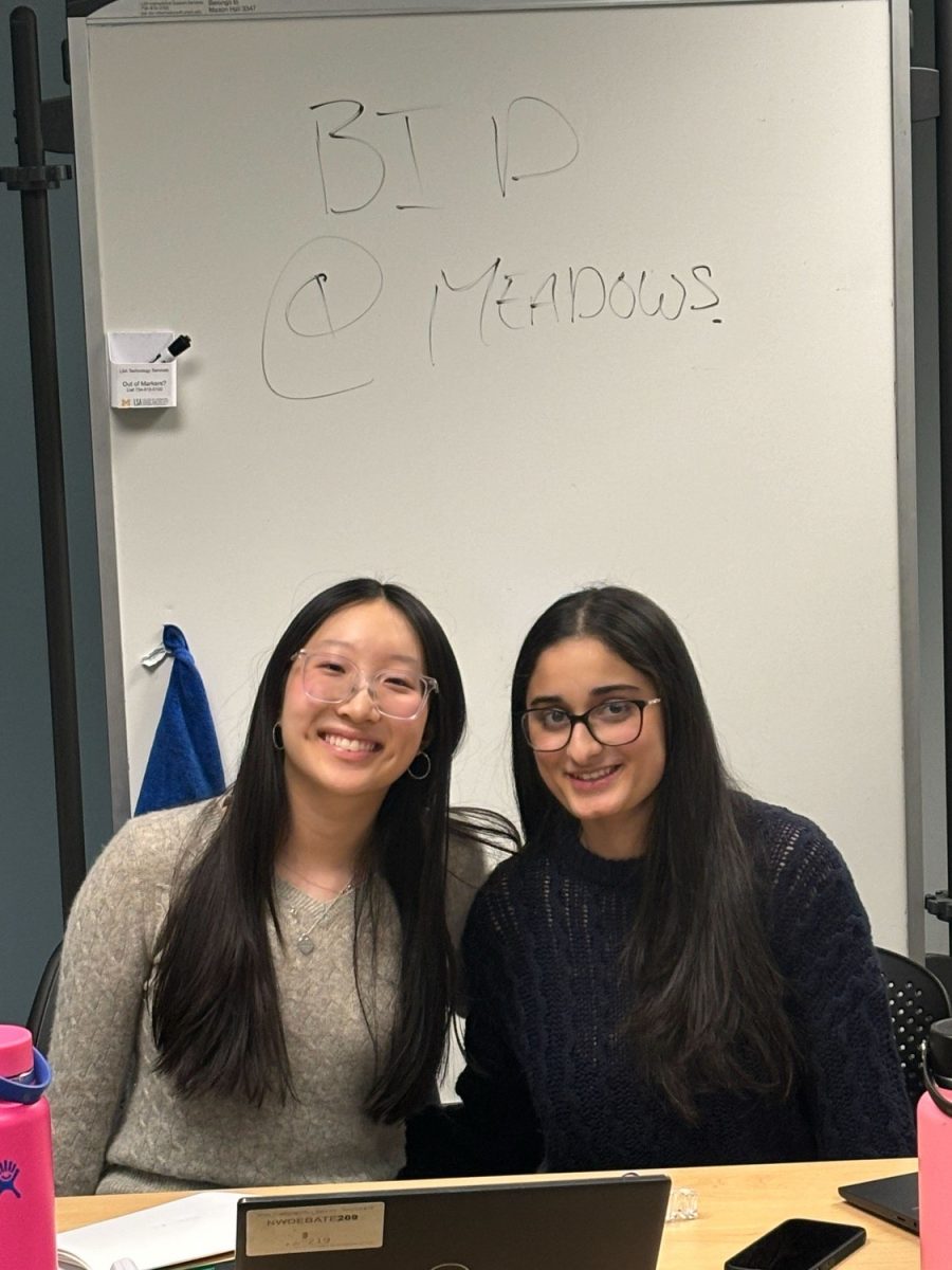 From left, juniors Ellie Kim and Ammara Vadiwala sit together in front of a sign showing off their victory of their first bid at Meadows. 