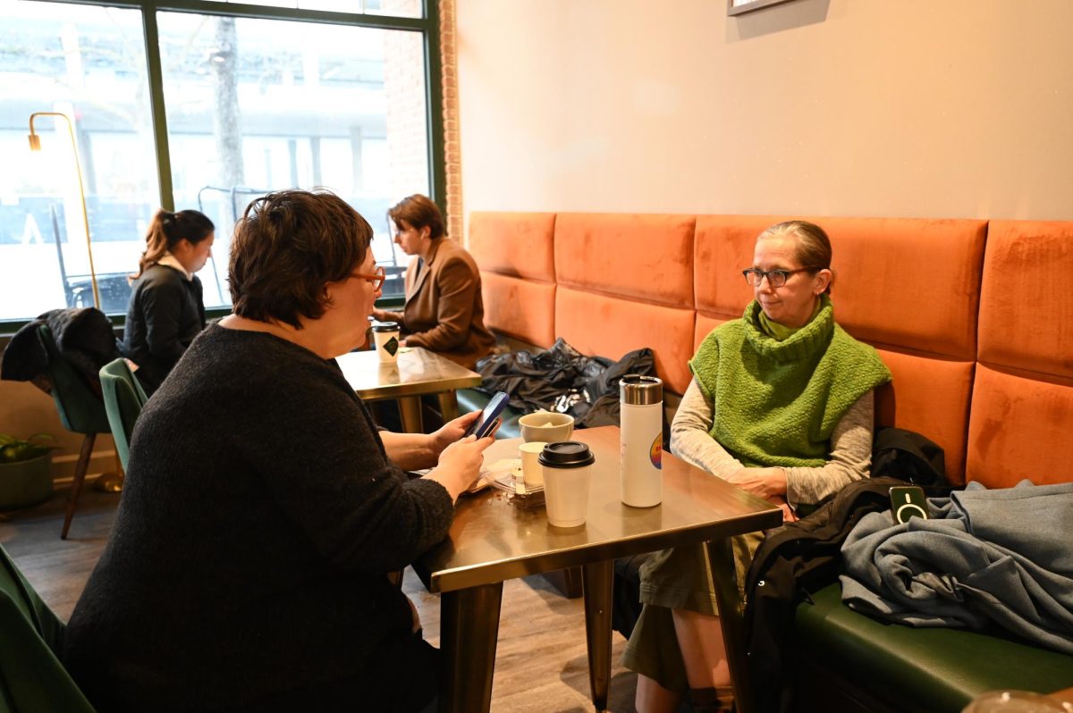 Two women sip their drinks, one using the “Start your day in Skokie” cup. They enjoy the opportunity to meet others that the environment of the cafe provides.